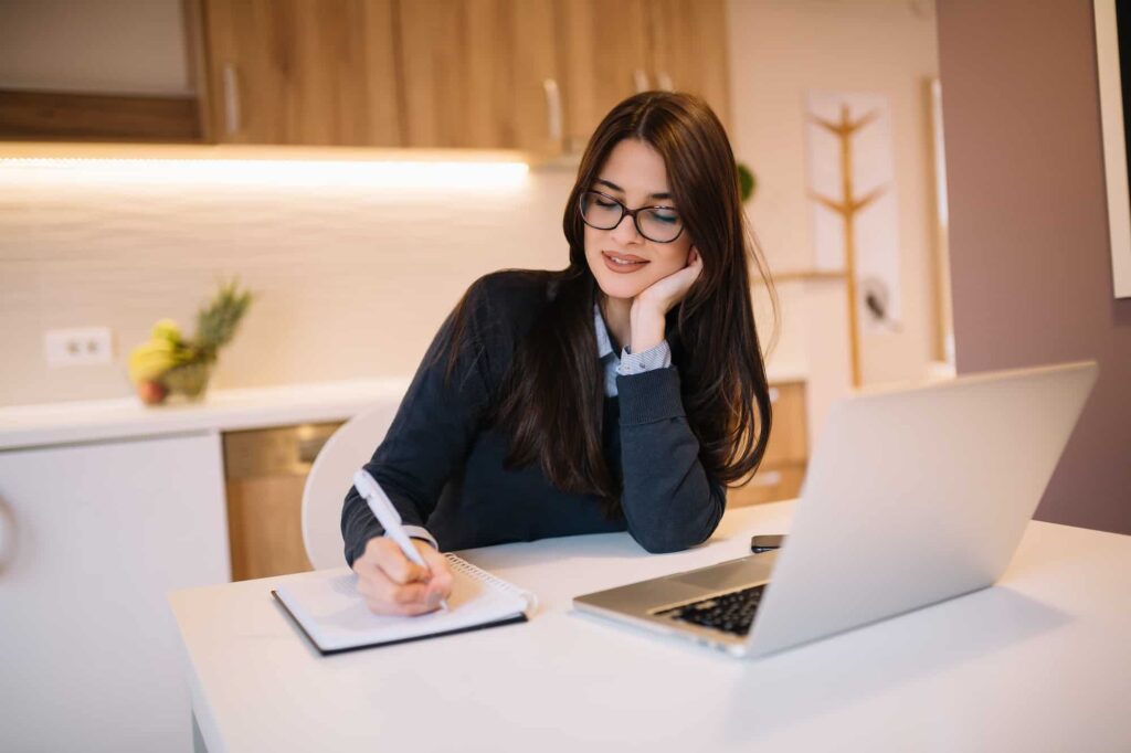 Woman writing in notebook with laptop in kitchen