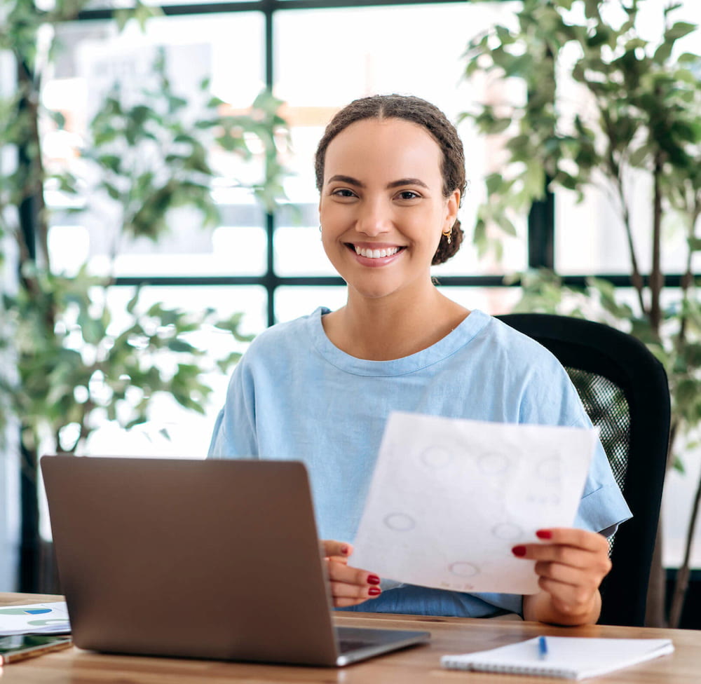 Smiling woman with document sitting at laptop desk.