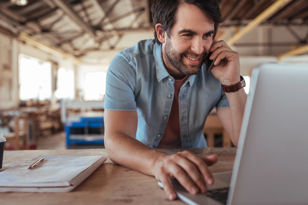 A man in a casual shirt talks on a smartphone while using a laptop in a well-lit workspace. Papers and a pen are on the table beside him, hinting at his focus on work or study, possibly reviewing WordPress maintenance plans.