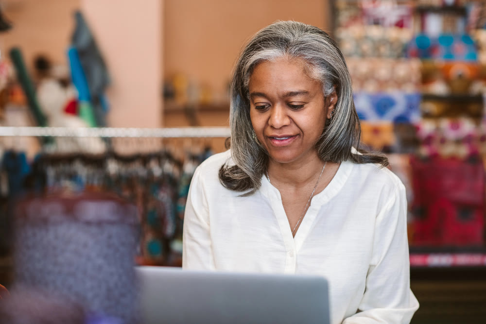 A woman with gray hair and a white blouse is sitting indoors, focused on her laptop. The colorful background suggests a retail or home setting, where she is engaged in updating wordpress maintenance plans.