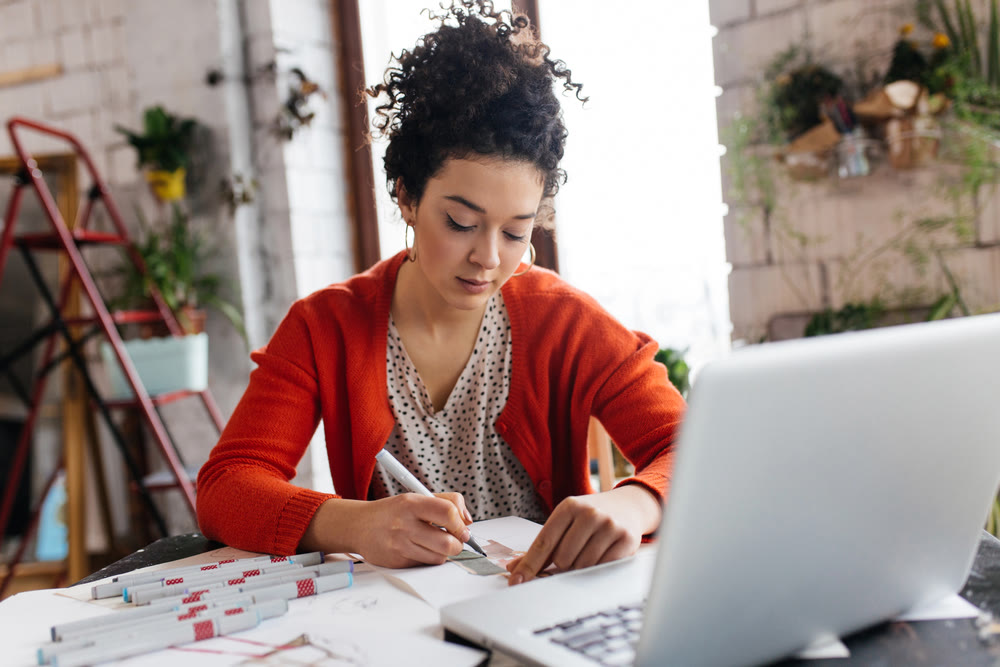 A woman with curly hair wearing a red cardigan is sitting at a desk, engrossed in sketching as she checks her laptop. Surrounded by art supplies, and plants on nearby shelves, she's researching WordPress maintenance plans for her creative projects.