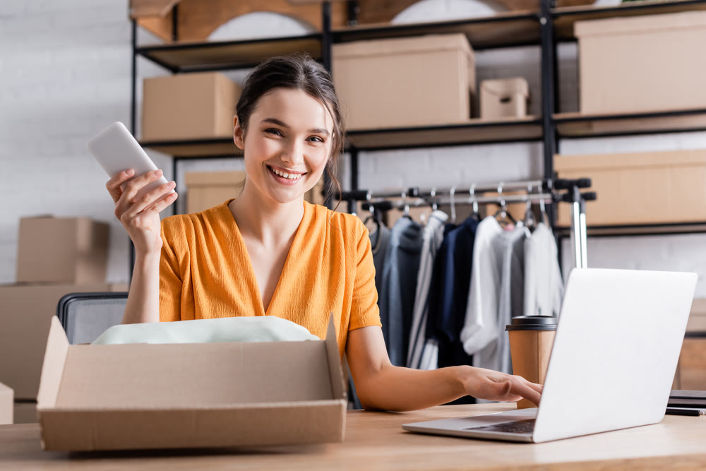 A woman in an orange shirt holds a smartphone and sits at a desk with a laptop, smiling amidst cardboard boxes and clothing racks. The scene suggests a small business or online shop setting where she could benefit from efficient WordPress maintenance plans to keep her digital storefront running smoothly.