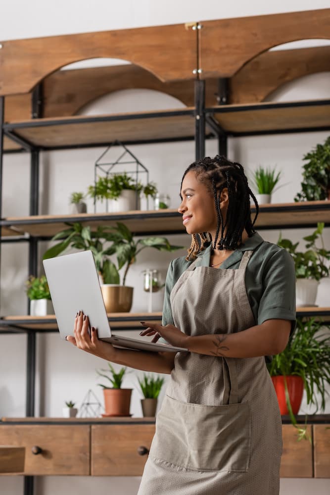 A woman wearing an apron is holding a laptop and smiling, standing in front of shelves filled with various potted plants. She has braided hair and appears to be in a greenhouse or plant shop, preparing to launch her business online with Movin on Up - The One Week Website.