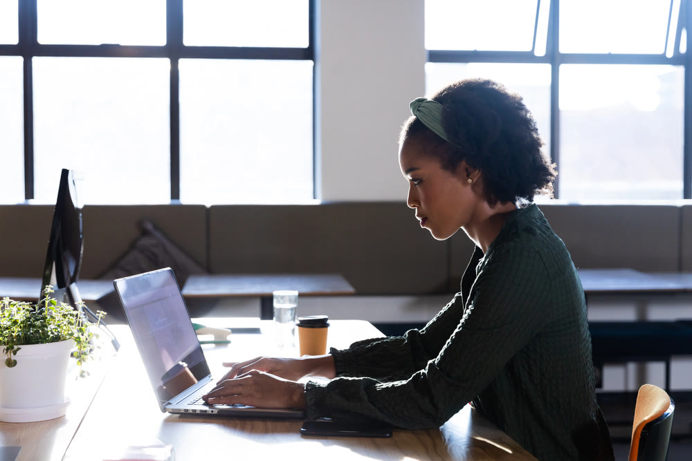 A woman sits at a desk working on her laptop in a well-lit office, meticulously reviewing WordPress maintenance plans. She is focused, with a potted plant, coffee cup, and water glass on the table. Large windows in the background let natural light fill the space.