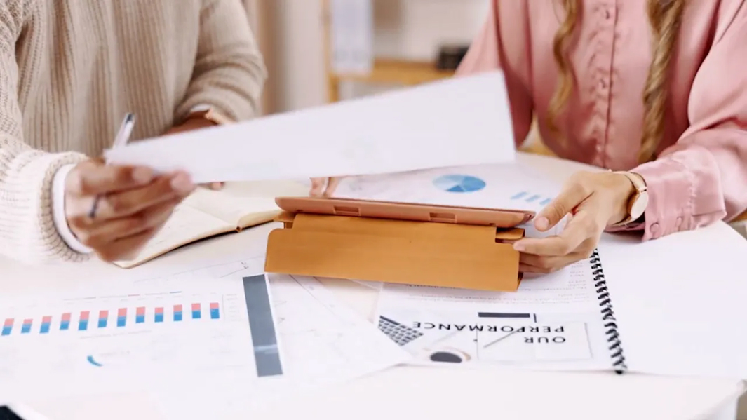Two people sit at a desk reviewing financial documents and charts. One holds a paper, while the other uses a tablet with a brown case. Among the paperwork and spiral-bound notebook, they discuss ways to improve website visitor retention.